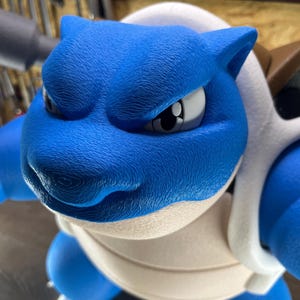 May include: A blue and white figurine of a turtle-like creature with a stern expression. The figure has a textured surface and is posed in a forward-facing position. The creature has a white shell and blue limbs.