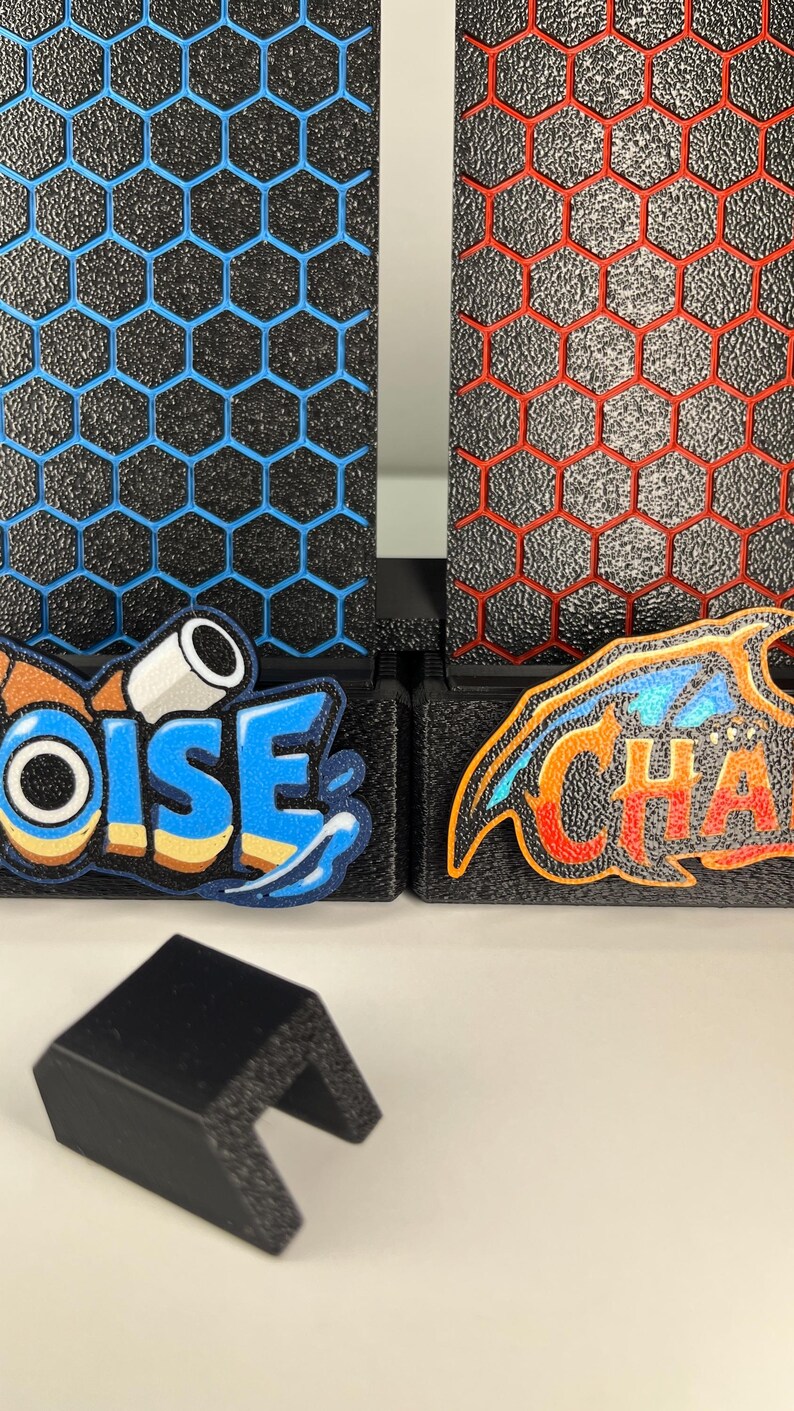 May include: Two black textured panels with a hexagonal grid pattern, one featuring blue outlines and the other red outlines. The panels display colorful embroidered patches with the text OISE and CHA. A small black U-shaped plastic clip sits in front.