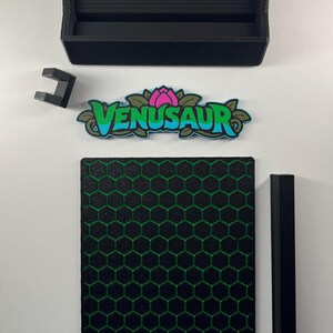 May include: A black rectangular tray, a small black bracket, a Venusaur nameplate with a pink flower and blue text, a square black panel with a green honeycomb pattern, and a long black rectangular bar arranged on a white surface.