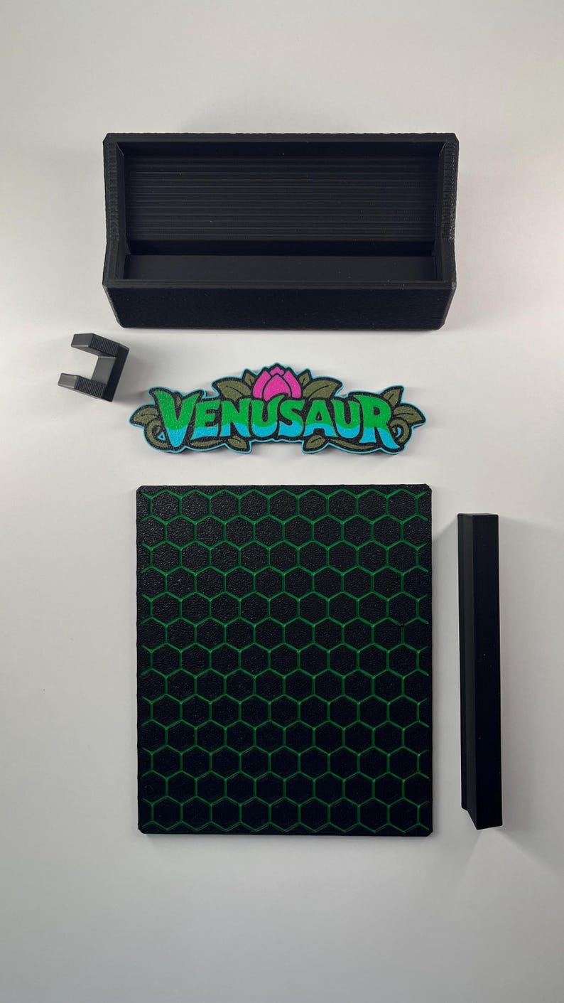 May include: A black rectangular tray, a small black bracket, a Venusaur nameplate with a pink flower and blue text, a square black panel with a green honeycomb pattern, and a long black rectangular bar arranged on a white surface.