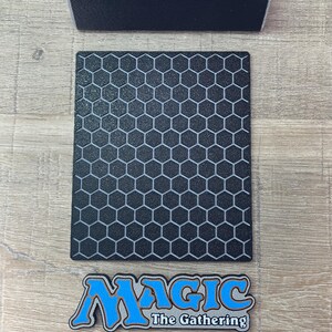 Op de afbeelding: Een zwarte rechthoekige kaartenstandaard met een gestructureerd honingraatpatroon op het voorpaneel. Onder het paneel bevindt zich een apart logoplaatje met de tekst Magic The Gathering in blauwe en zilveren letters. Een zwarte basis is inbegrepen.