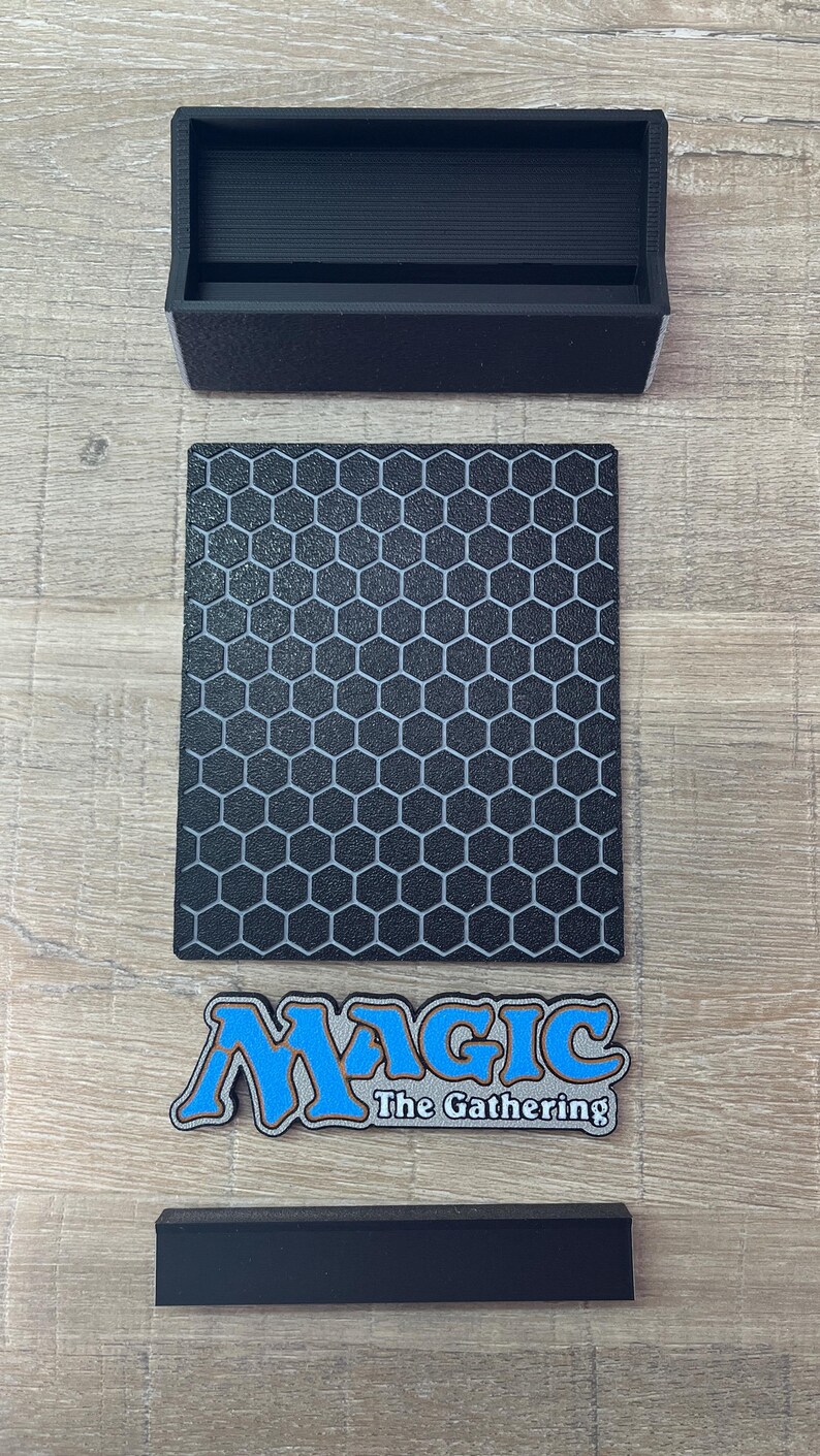 Op de afbeelding: Een zwarte rechthoekige kaartenstandaard met een gestructureerd honingraatpatroon op het voorpaneel. Onder het paneel bevindt zich een apart logoplaatje met de tekst Magic The Gathering in blauwe en zilveren letters. Een zwarte basis is inbegrepen.