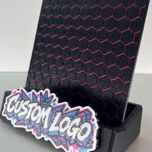K&ouml;nnte beinhalten: Ein schwarzer Pr&auml;sentationsst&auml;nder mit texturiertem Wabenmuster und magentafarbenen Akzenten. Die Vorderseite des Sockels zeigt eine wei&szlig;-blau-violette Grafik im Graffiti-Stil mit dem Text CUSTOM LOGO in fetten, wei&szlig;en Gro&szlig;buchstaben.