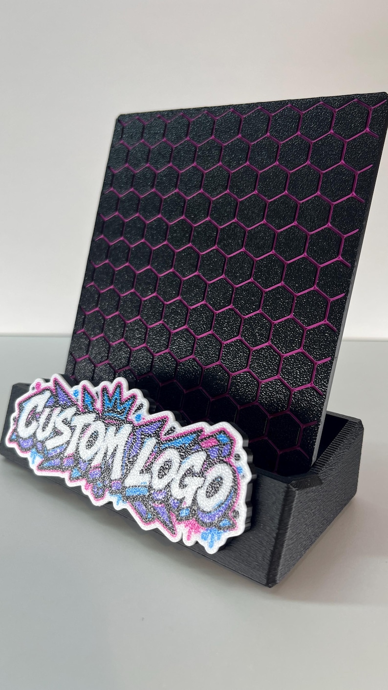 K&ouml;nnte beinhalten: Ein schwarzer Pr&auml;sentationsst&auml;nder mit texturiertem Wabenmuster und magentafarbenen Akzenten. Die Vorderseite des Sockels zeigt eine wei&szlig;-blau-violette Grafik im Graffiti-Stil mit dem Text CUSTOM LOGO in fetten, wei&szlig;en Gro&szlig;buchstaben.