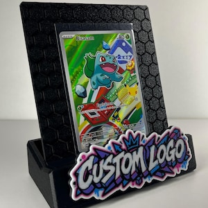 K&ouml;nnte beinhalten: Ein schwarzer Aufsteller mit sechseckigem Strukturmuster, der eine Sammelkarte mit der Figur Bisasam h&auml;lt. Auf der Vorderseite befindet sich ein wei&szlig;-lila-blaues Graffiti-Schild mit der Aufschrift CUSTOM LOGO.
