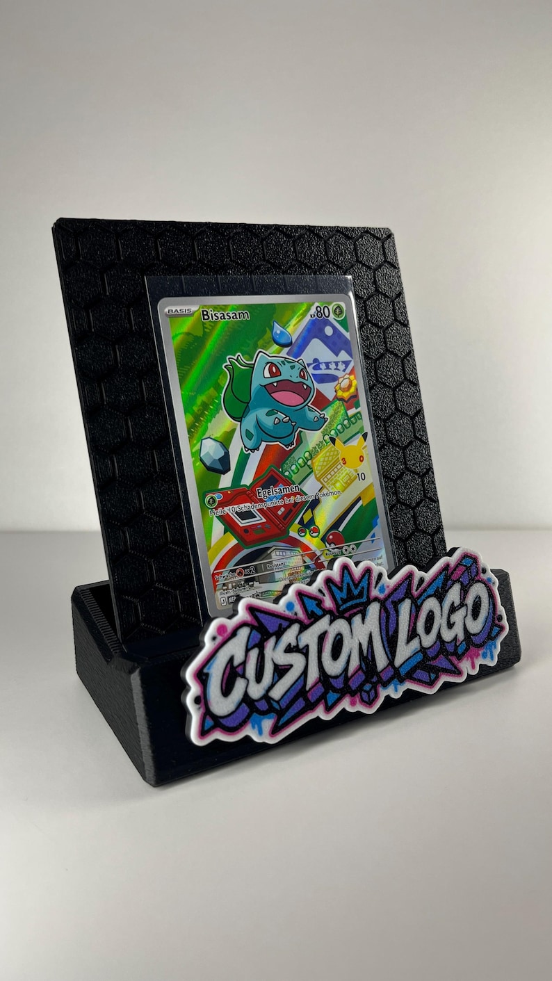 K&ouml;nnte beinhalten: Ein schwarzer Aufsteller mit sechseckigem Strukturmuster, der eine Sammelkarte mit der Figur Bisasam h&auml;lt. Auf der Vorderseite befindet sich ein wei&szlig;-lila-blaues Graffiti-Schild mit der Aufschrift CUSTOM LOGO.