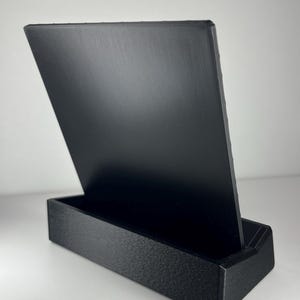 K&ouml;nnte beinhalten: Ein schwarzer, rechteckiger Pr&auml;sentationsst&auml;nder mit einer strukturierten Basis und einer glatten, aufrechten R&uuml;ckwand. Das Objekt besteht aus einem soliden, mattschwarzen Material mit sichtbaren Schichtlinien aus dem Fertigungsprozess.