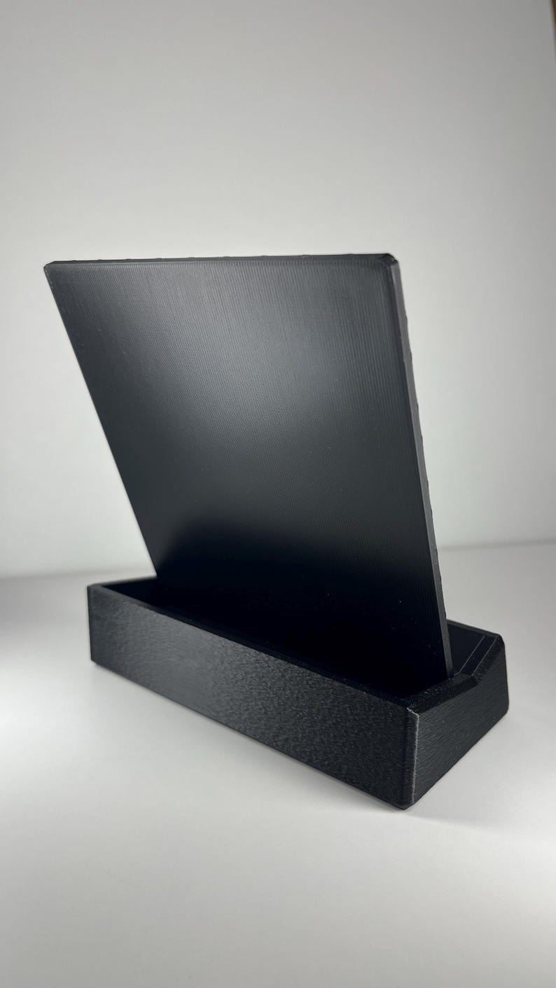 K&ouml;nnte beinhalten: Ein schwarzer, rechteckiger Pr&auml;sentationsst&auml;nder mit einer strukturierten Basis und einer glatten, aufrechten R&uuml;ckwand. Das Objekt besteht aus einem soliden, mattschwarzen Material mit sichtbaren Schichtlinien aus dem Fertigungsprozess.