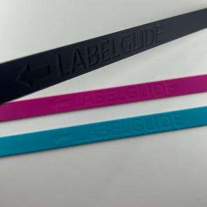Peut inclure: Trois bandes fines, plates et flexibles de couleur noire, magenta et bleu sarcelle. Chaque bande pr&eacute;sente le texte en relief LABELGLIDE et une ic&ocirc;ne de fl&egrave;che pointant vers la gauche. Les bandes sont dispos&eacute;es en diagonale.