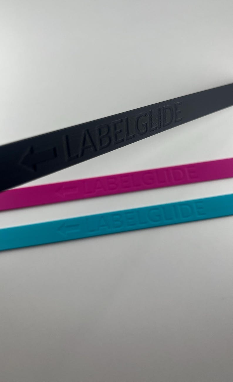 Peut inclure: Trois bandes fines, plates et flexibles de couleur noire, magenta et bleu sarcelle. Chaque bande pr&eacute;sente le texte en relief LABELGLIDE et une ic&ocirc;ne de fl&egrave;che pointant vers la gauche. Les bandes sont dispos&eacute;es en diagonale.