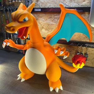 May include: A vibrant orange Charizard figurine with blue wings and white claws. The figurine has a cream-colored belly and is holding a red and yellow flame. The figure is a popular character from the Pokemon franchise.