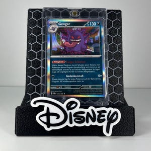 May include: A Gengar Pokemon trading card in a clear protective case, displayed on a black hexagonal-patterned stand with the white Disney logo text at the base. The card features a purple ghost character with a long tongue and 130 HP.