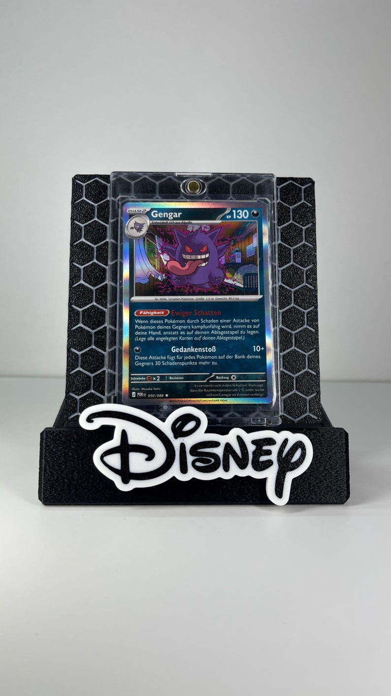 May include: A Gengar Pokemon trading card in a clear protective case, displayed on a black hexagonal-patterned stand with the white Disney logo text at the base. The card features a purple ghost character with a long tongue and 130 HP.