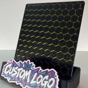K&ouml;nnte beinhalten: Ein schwarzer Aufsteller mit einer vertikalen Platte mit gelbem Wabenmuster. An der Vorderseite befindet sich ein wei&szlig;es Schild mit dem Text CUSTOM LOGO in stilisierter Schrift, verziert mit blauen, rosa und violetten Glitzerakzenten.