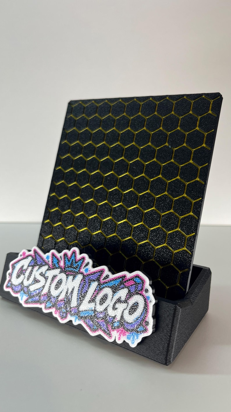 K&ouml;nnte beinhalten: Ein schwarzer Aufsteller mit einer vertikalen Platte mit gelbem Wabenmuster. An der Vorderseite befindet sich ein wei&szlig;es Schild mit dem Text CUSTOM LOGO in stilisierter Schrift, verziert mit blauen, rosa und violetten Glitzerakzenten.