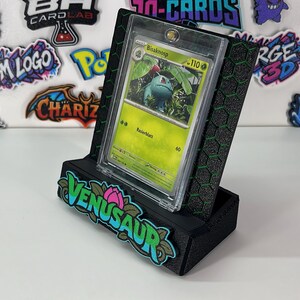 May include: A black display stand with a green honeycomb pattern holds a clear protective case containing a green Bulbasaur trading card. The front of the stand features a colorful, raised logo that reads VENUSAUR with a pink flower icon.