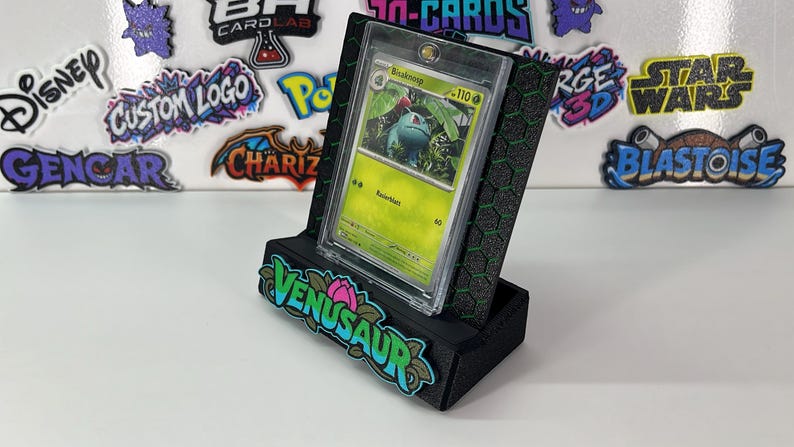 May include: A black display stand with a green honeycomb pattern holds a clear protective case containing a green Bulbasaur trading card. The front of the stand features a colorful, raised logo that reads VENUSAUR with a pink flower icon.