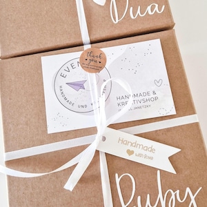 May include: Two brown cardboard gift boxes tied with white ribbon. Each box features a white tag reading Handmade with love and a white name decal, Dua and Ruby. A circular thank you sticker and a white business card with a purple paper plane.