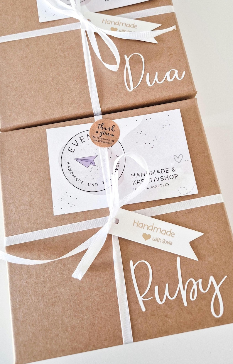 May include: Two brown cardboard gift boxes tied with white ribbon. Each box features a white tag reading Handmade with love and a white name decal, Dua and Ruby. A circular thank you sticker and a white business card with a purple paper plane.