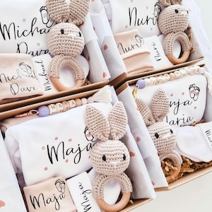 May include: Four gift boxes containing personalized baby items including white fabric pieces with names like Michael Dave, Nuri, Maja, and Ninja Maria, crochet bunny rattles with wooden rings, and beaded pacifier clips with lettered beads.