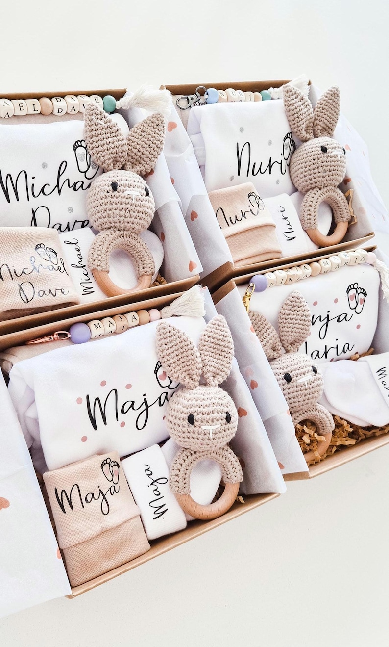 May include: Four gift boxes containing personalized baby items including white fabric pieces with names like Michael Dave, Nuri, Maja, and Ninja Maria, crochet bunny rattles with wooden rings, and beaded pacifier clips with lettered beads.