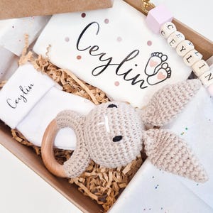 May include: A gift box containing a white baby bodysuit with the name Ceylin and baby feet printed on it, a white sock with Ceylin printed on it, a beige crocheted rabbit rattle with a wooden ring, and a keychain with the name Ceylin in beads.