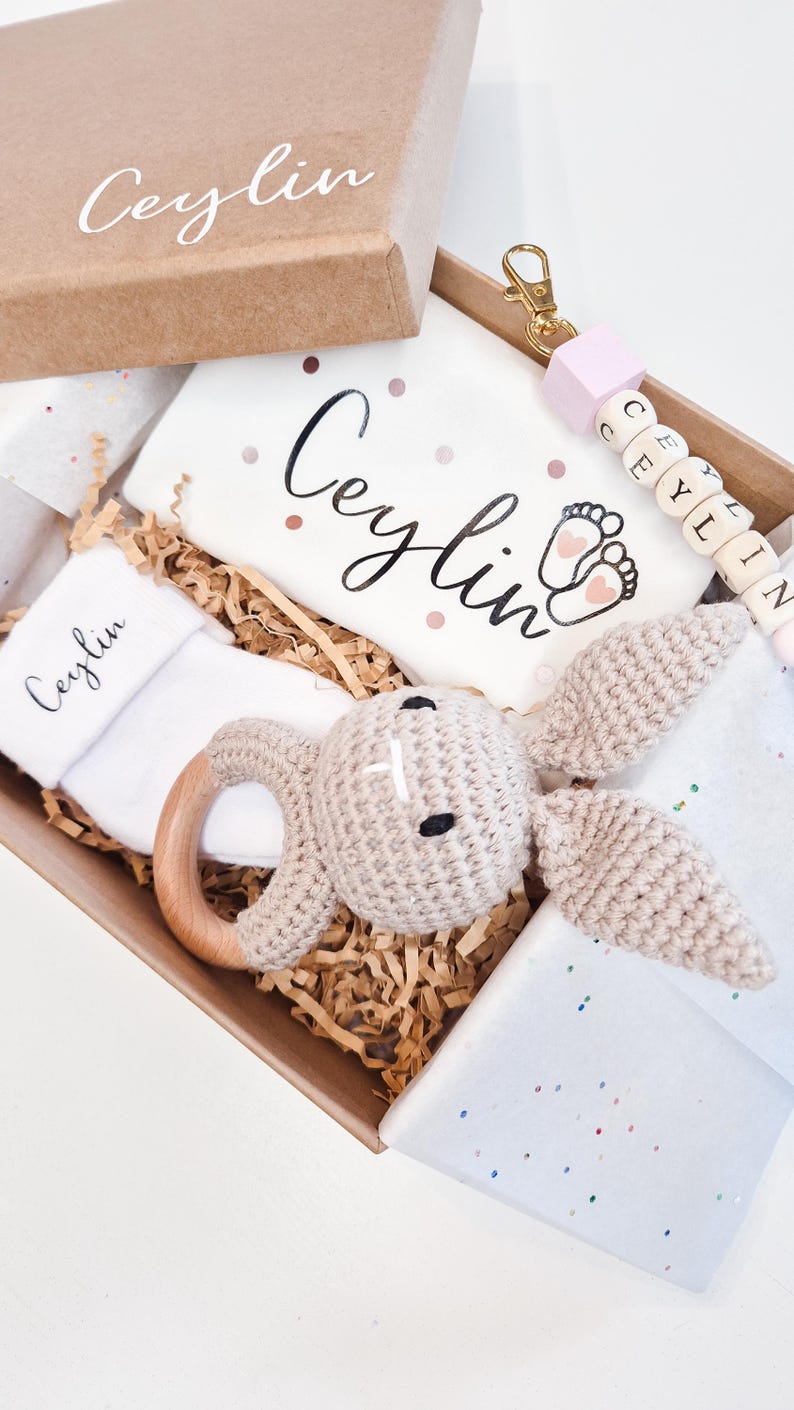 May include: A gift box containing a white baby bodysuit with the name Ceylin and baby feet printed on it, a white sock with Ceylin printed on it, a beige crocheted rabbit rattle with a wooden ring, and a keychain with the name Ceylin in beads.