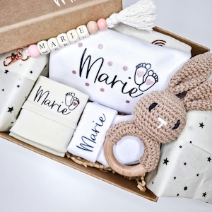 May include: A cardboard gift box containing a white baby bodysuit with the name Marie and footprint graphics, a crochet bunny rattle with a wooden ring, a beaded keychain with the name Marie, and folded white fabric items with the name Marie.