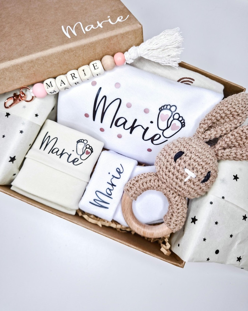 May include: A cardboard gift box containing a white baby bodysuit with the name Marie and footprint graphics, a crochet bunny rattle with a wooden ring, a beaded keychain with the name Marie, and folded white fabric items with the name Marie.