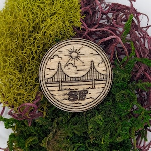 Puede incluir: Un emblema redondo de madera con el puente Golden Gate, el sol, las nubes y las letras "SF". El emblema está rodeado de musgos verdes, verde amarillentos y granates.