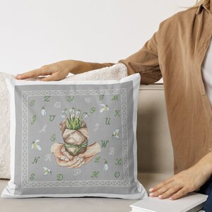 Puede incluir: Una almohada gris con un borde blanco y un diseño de punto de cruz que presenta un ramo de campanillas de invierno en un paquete de papel marrón sostenido por dos manos. El diseño está rodeado por un círculo de letras del alfabeto.