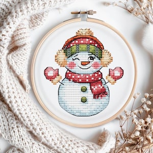 Puede incluir: Bordado de punto de cruz de un muñeco de nieve guiñando un ojo, con gorro, orejeras, bufanda y mitones rojos y verdes. El muñeco de nieve está bordado sobre tela blanca, en un bastidor de bordado de madera. El diseño tiene un tema de vacaciones de invierno.