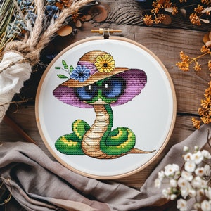 Puede incluir: Una serpiente verde que lleva un sombrero de paja con ribete rosa y gafas de sol. El sombrero tiene una flor amarilla y una flor azul.