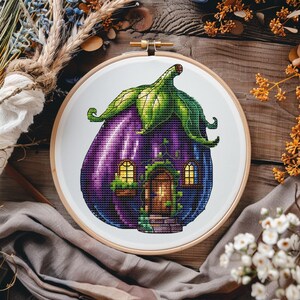 Puede incluir: Un aro de bordado de punto de cruz con una casa con forma de berenjena morada, un techo verde, una puerta marrón y dos ventanas con luces amarillas. La casa está rodeada de enredaderas verdes.