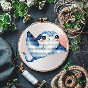 Puede incluir: Un bastidor de bordado de punto de cruz con un diseño de manta raya azul sonriente. La obra de arte está sobre un fondo degradado rosa suave. El bastidor está rodeado de materiales de artesanía y vegetación.