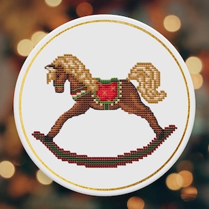 Puede incluir: Adorno navideño redondo y blanco con borde dorado. Presenta un diseño de punto de cruz de un caballo balancín marrón con crin y cola rubias, y una silla de montar roja y verde. El caballo balancín está sobre una base verde y roja.