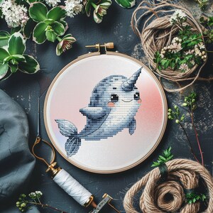 Puede incluir: Un aro de bordado redondo con un fondo blanco y un degradado rosa. Un narval de dibujos animados azul y gris con un cuerno rosa y ojos grandes está bordado en el aro.