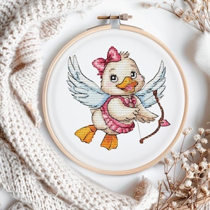 Puede incluir: Bordado de punto de cruz de un patito vestido de Cupido, sosteniendo un arco y una flecha. El patito tiene plumas blancas, un lazo rosa y un atuendo rosa. El bordado está en un aro de madera, con fondo blanco.