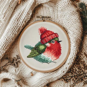Puede incluir: Un aro de bordado de punto de cruz con un pájaro rojo que lleva un gorro de punto rojo y blanco con un pompón blanco. El pájaro tiene alas verdes y una barriga blanca.