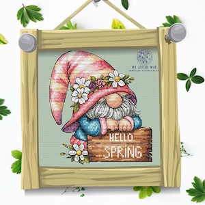 Puede incluir: Arte de punto de cruz de un gnomo de jardín con un sombrero rosa adornado con margaritas blancas y un atuendo azul. El gnomo sostiene un letrero de madera que dice "HELLO SPRING". La obra de arte está enmarcada en un marco de madera marrón claro.