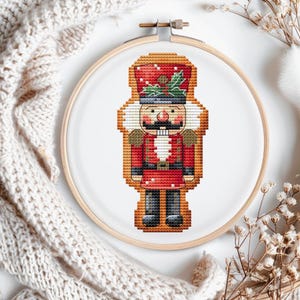 May include: A cross-stitch pattern of a nutcracker in a red and white uniform with a gold belt and a red hat with white polka dots and green holly.