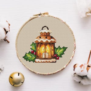 Puede incluir: Bordado de punto de cruz de una linterna marrón con una vela encendida en su interior, rodeada de hojas verdes de acebo y bayas rojas. El diseño está en un bastidor de bordado redondo de madera. Una campana dorada y acentos de algodón están en el fondo.