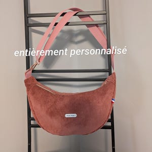 May include: A crescent-shaped dusty rose corduroy crossbody bag with a wide pink adjustable strap, a top zipper closure, and a small white label reading Hand Made. The text entièrement personnalisé is visible.