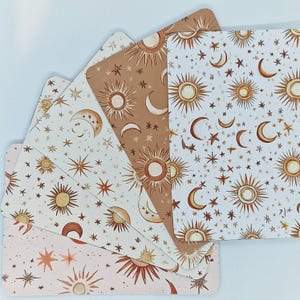 Puede incluir: Un conjunto de cartas de tarot con diseños celestiales. Cada carta presenta un patrón diferente de soles, lunas y estrellas en tonos marrones, beige y rosas. Las cartas están dispuestas en forma de abanico, mostrando la variedad de ilustraciones.