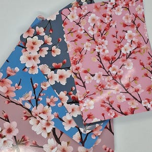 Op de afbeelding: Vier rechthoekige notitieboekjes met een bloemig kersenbloesemdesign. De notitieboekjes hebben roze, blauwe en grijze achtergronden met takken van roze en witte bloemen. De notitieboekjes zijn op elkaar gestapeld.