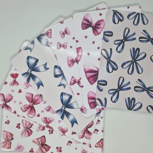 May include: A selection of decorative cards with bow patterns. The cards showcase designs with pink and blue bows, small hearts, and floral details. The cards have a rounded top edge and a white background.