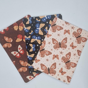Op de afbeelding: Drie decoratieve rechthoekige notitieboekjes met vlinderpatronen. Eén is bruin met rode en witte vlinders, een andere is zwart met blauwe en gouden vlinders, en de derde is beige met bruine vlinders.