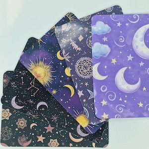 Può includere: Un set di carte dei tarocchi con disegni celesti. Ogni carta presenta un colore di sfondo diverso, tra cui nero, viola e blu navy. Le carte sono decorate con lune, stelle, soli e altri elementi cosmici. Le carte sono a ventaglio, mostrando la varietà dei disegni.
