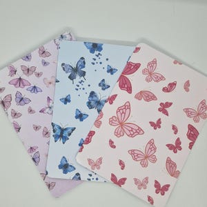 Op de afbeelding: Drie decoratieve notitieboekjes met vlindermotieven in roze, paarse en blauwe tinten. De vlinderillustraties zijn verspreid over de pagina's, wat een grillige en artistieke uitstraling creëert. Ideaal om te journalen of notities te maken.