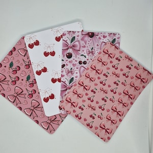 Op de afbeelding: Vier decoratieve kaarten met kersen- en strikpatronen. De kaarten hebben verschillende ontwerpen, waaronder kersen met strikken, strikken met harten en herhalende strik- en kersenpatronen in roze en witte tinten.